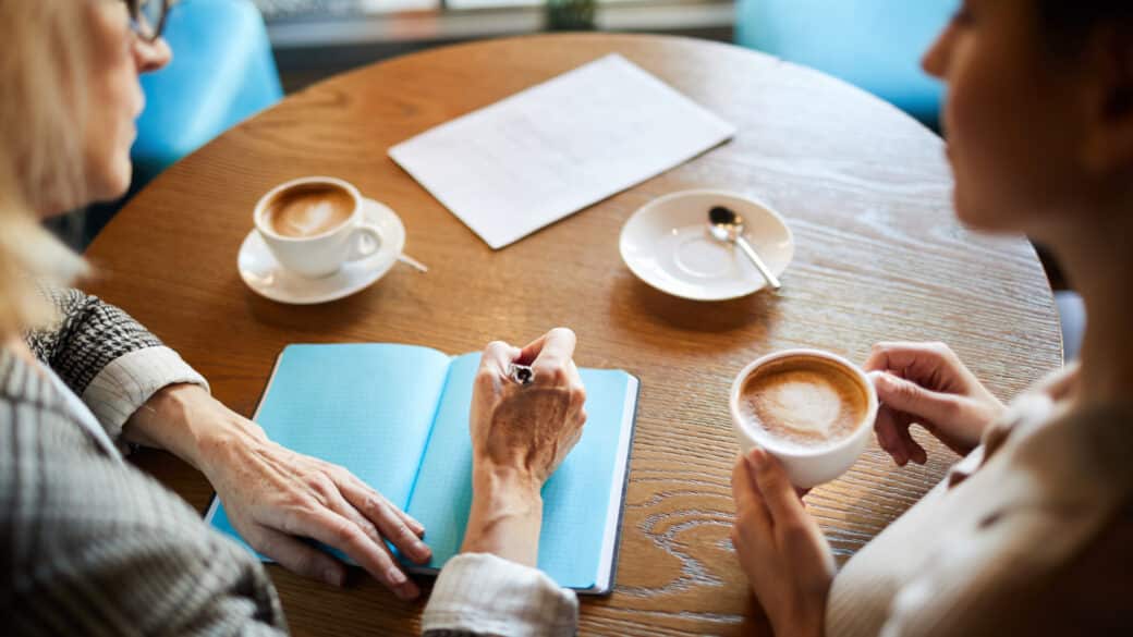 One of businesswomen making notes in notepad during discussion of organization moments with colleague by cup of cappuccino